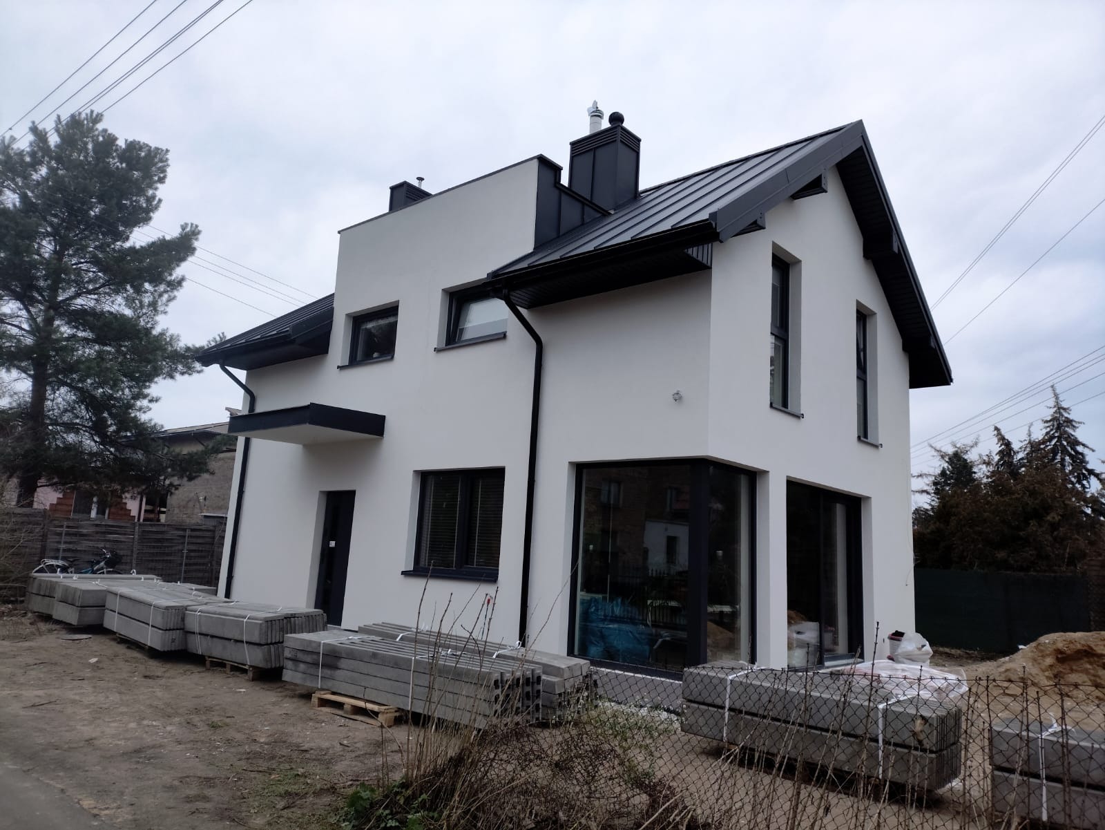 Nowoczesny, dwukondygnacyjny dom murowany z czarnym dachem blaszanym i grafitowymi oknami, z paletami szarych kostek brukowych ułożonymi wzdłuż ściany, teren wokół domu jeszcze niezagospodarowany.