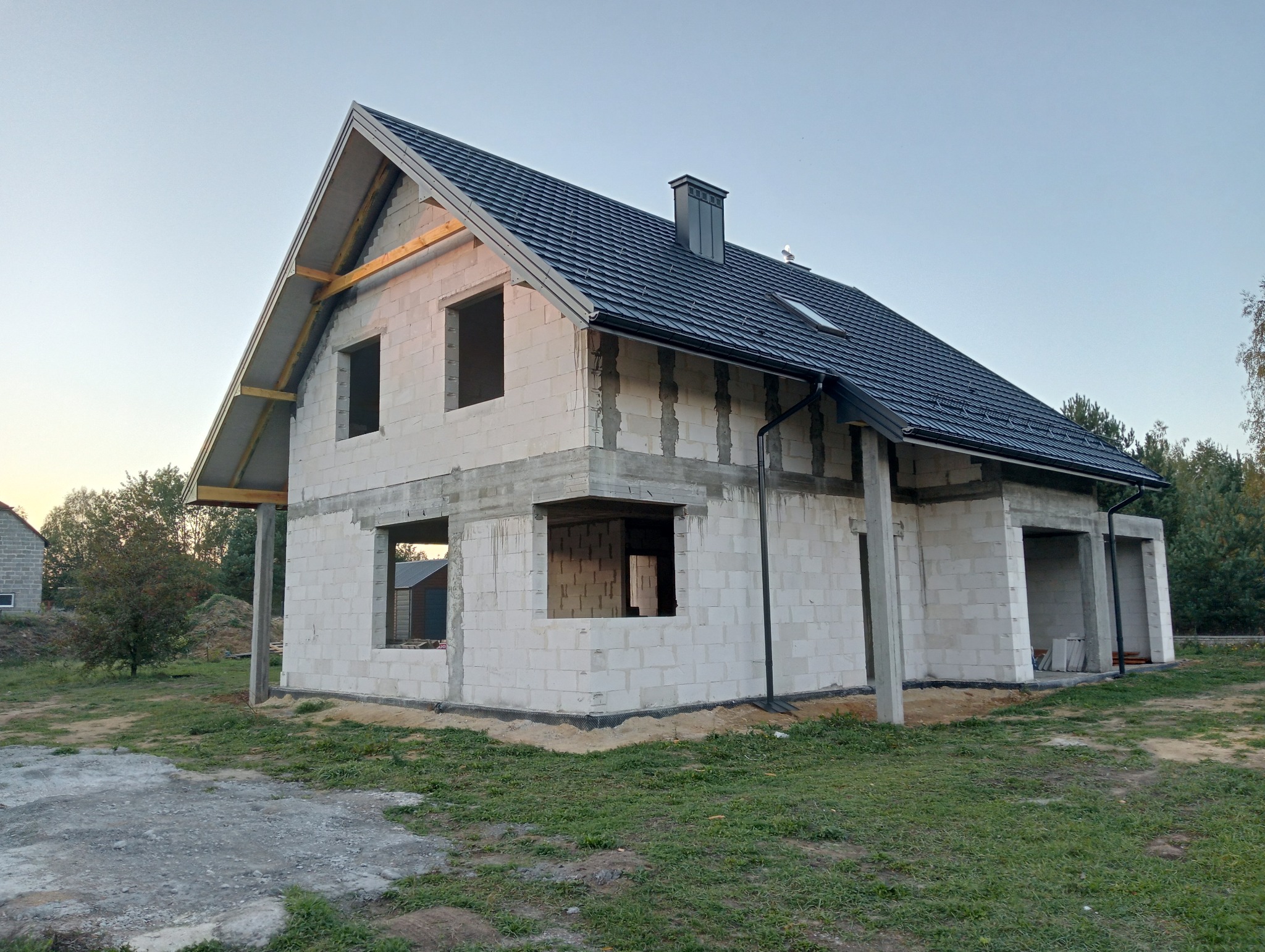 Niedokończony dwukondygnacyjny budynek z białych bloczków z ciemną dachówką, widoczne niezamaskowane betonowe słupy i brak okien, w tle drzewa i błękitne niebo.