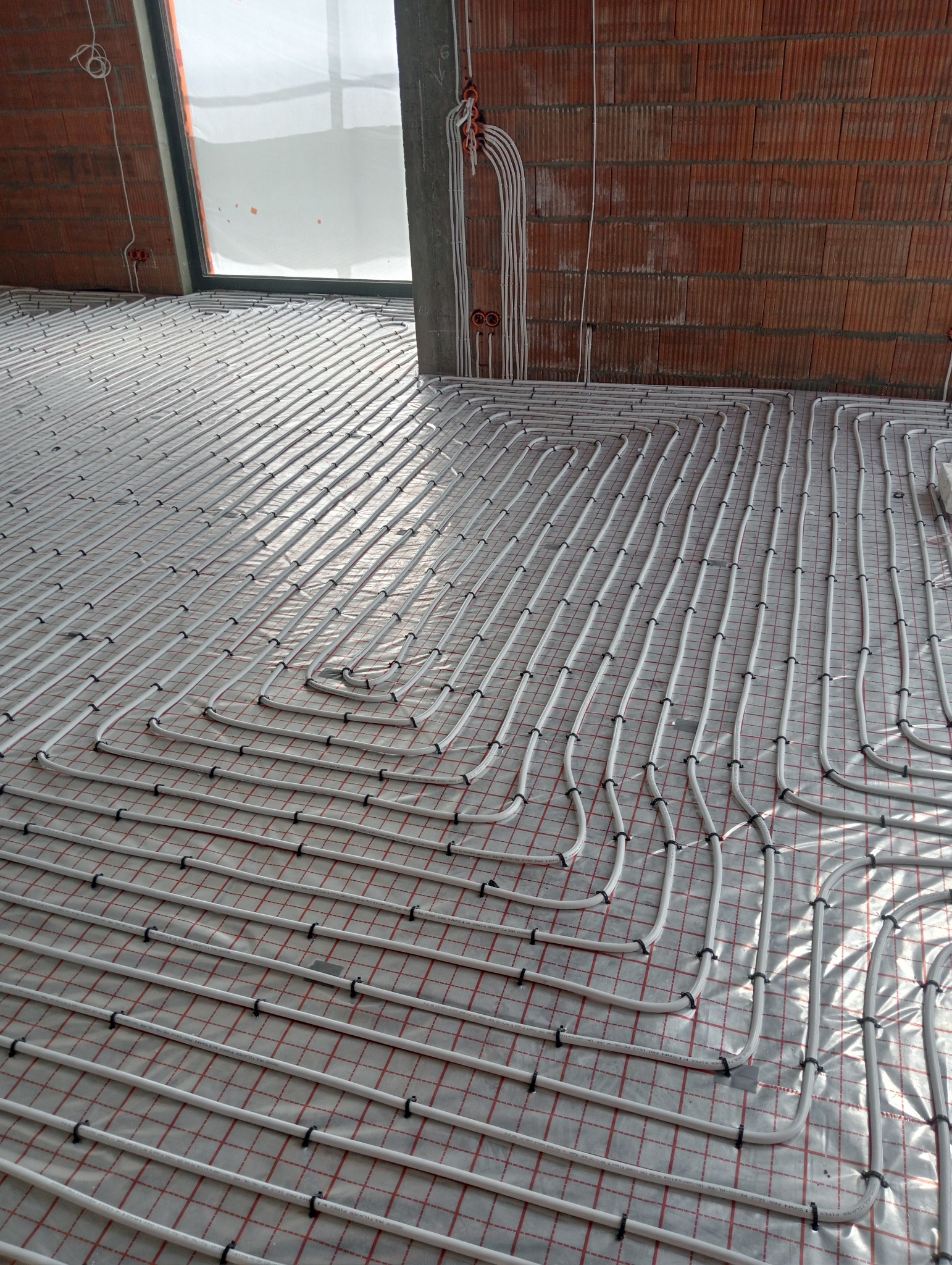 Instalacja ogrzewania podłogowego z białych rur na folii z czerwoną siatką w surowym stanie budynku, widoczne przewody elektryczne na ścianie z cegły.