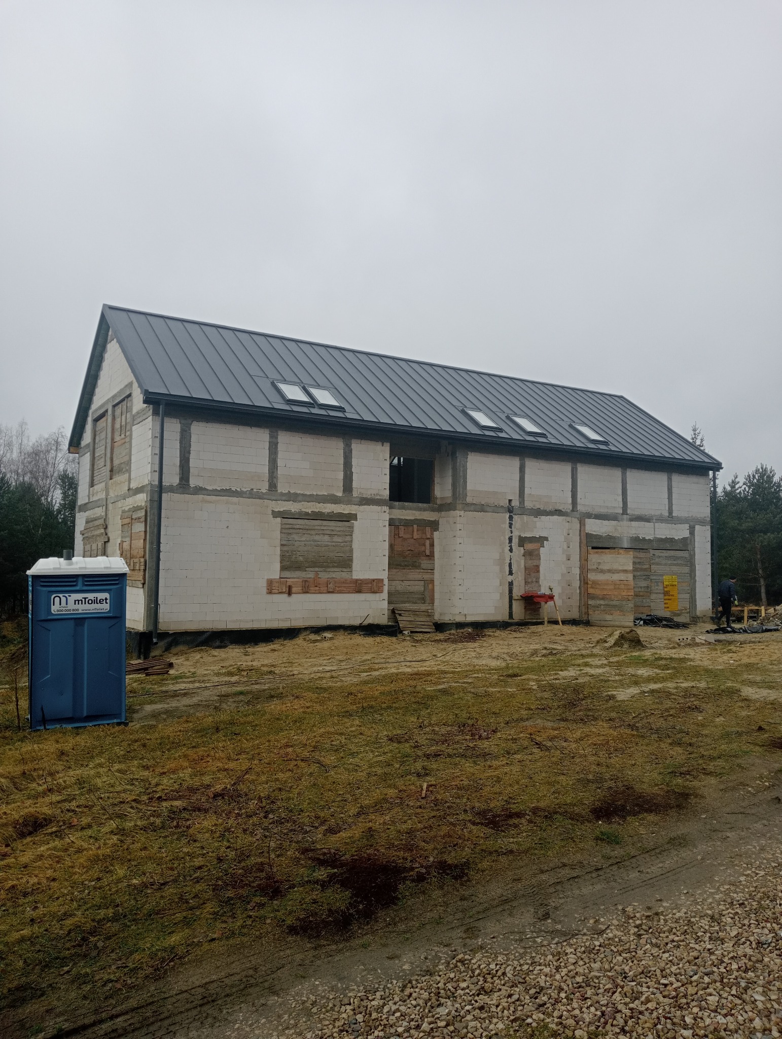 Dwukondygnacyjny budynek w stanie surowym zamkniętym z szarej cegły, z ciemnoszarym dachem z oknami dachowymi, zabezpieczone deskami otwory okienne, na zewnątrz przenośna toaleta.