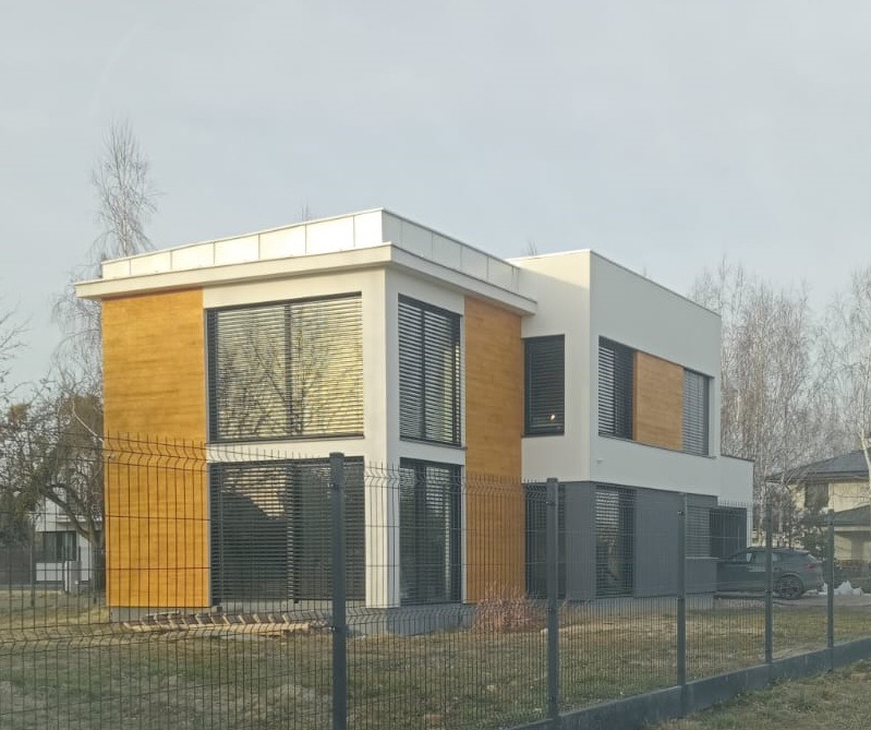Nowoczesny, dwukondygnacyjny dom z elementami drewnianej elewacji i dużymi oknami z żaluzjami, widziany zza metalowego ogrodzenia.
