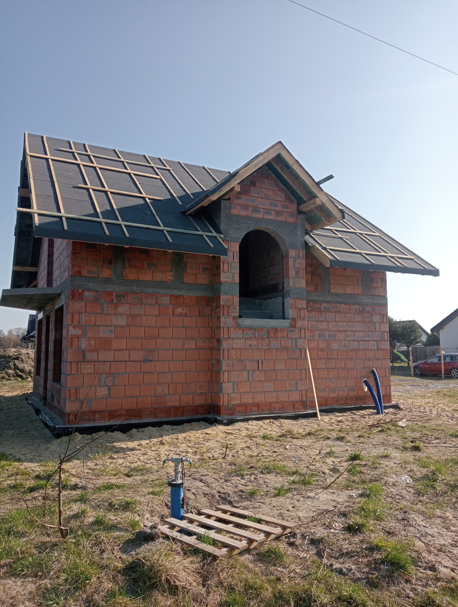 Surowy stan budynku z czerwonej cegły z widocznym zadaszeniem, oknem w kształcie łuku i nieotynkowanymi spoinami, na tle błękitnego nieba.