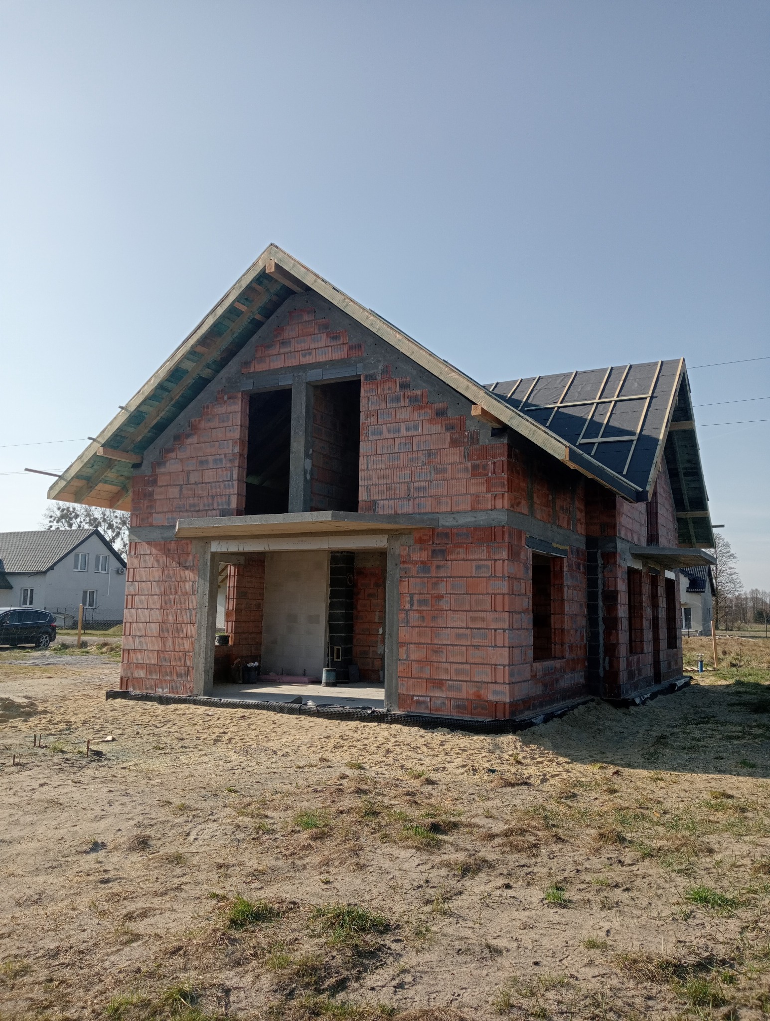 Surowy stan domu z czerwonej cegły, widoczne otwarte okna i drzwi, częściowo pokryty dach z czarną membraną, na pierwszym planie piaszczysta ziemia.