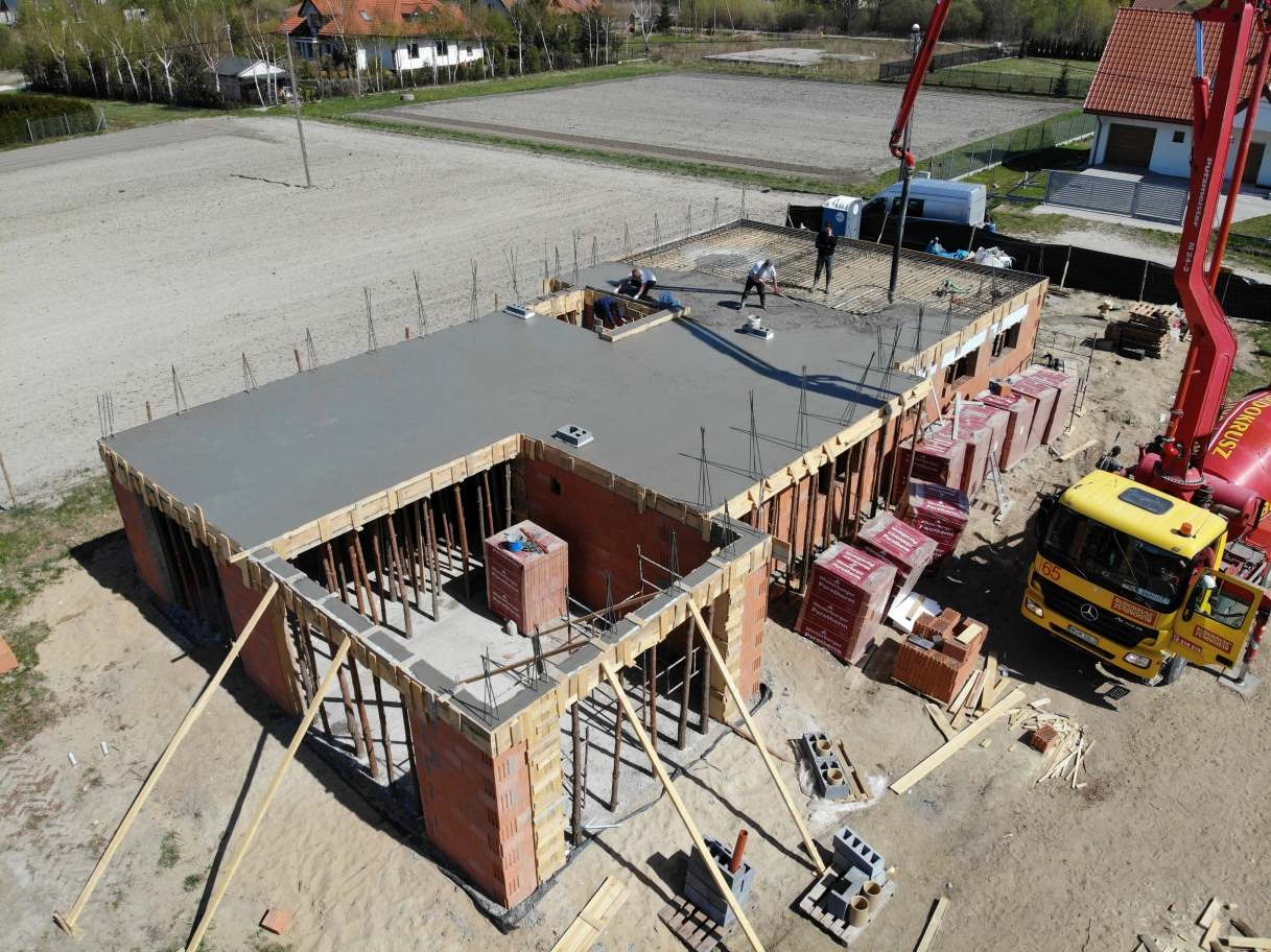 Wysoki kąt: Budowa domu murowanego z wylanym betonowym stropem, widoczne podpory szalunkowe, palety z cegłami Porotherm i pompa do betonu z żółtą ciężarówką w tle.