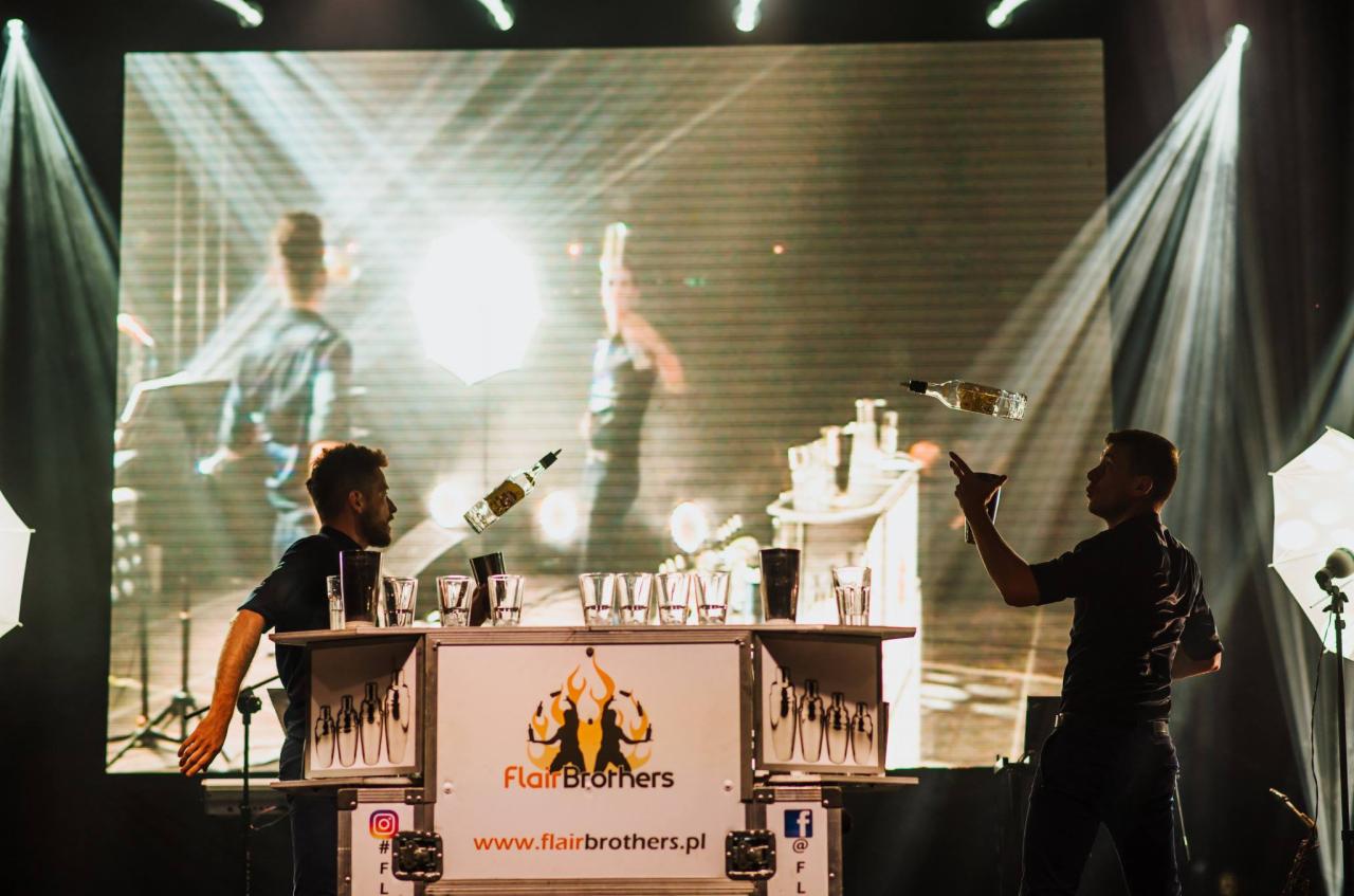 Barmani Flair Brothers w trakcie pokazu: jeden żongluje butelką, drugi czeka z wyciągniętą ręką, na stole rząd szklanek i logo firmy, w tle ekran z powtórką występu.