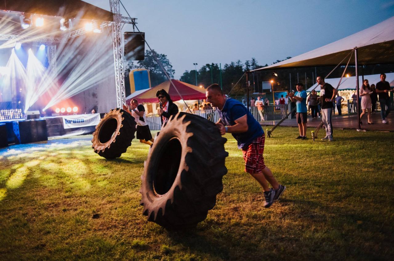 Mężczyzna w czerwonych szortach i niebieskiej koszulce przepycha ogromną oponę na trawie podczas eventu plenerowego, w tle scena oświetlona reflektorami i publiczność.