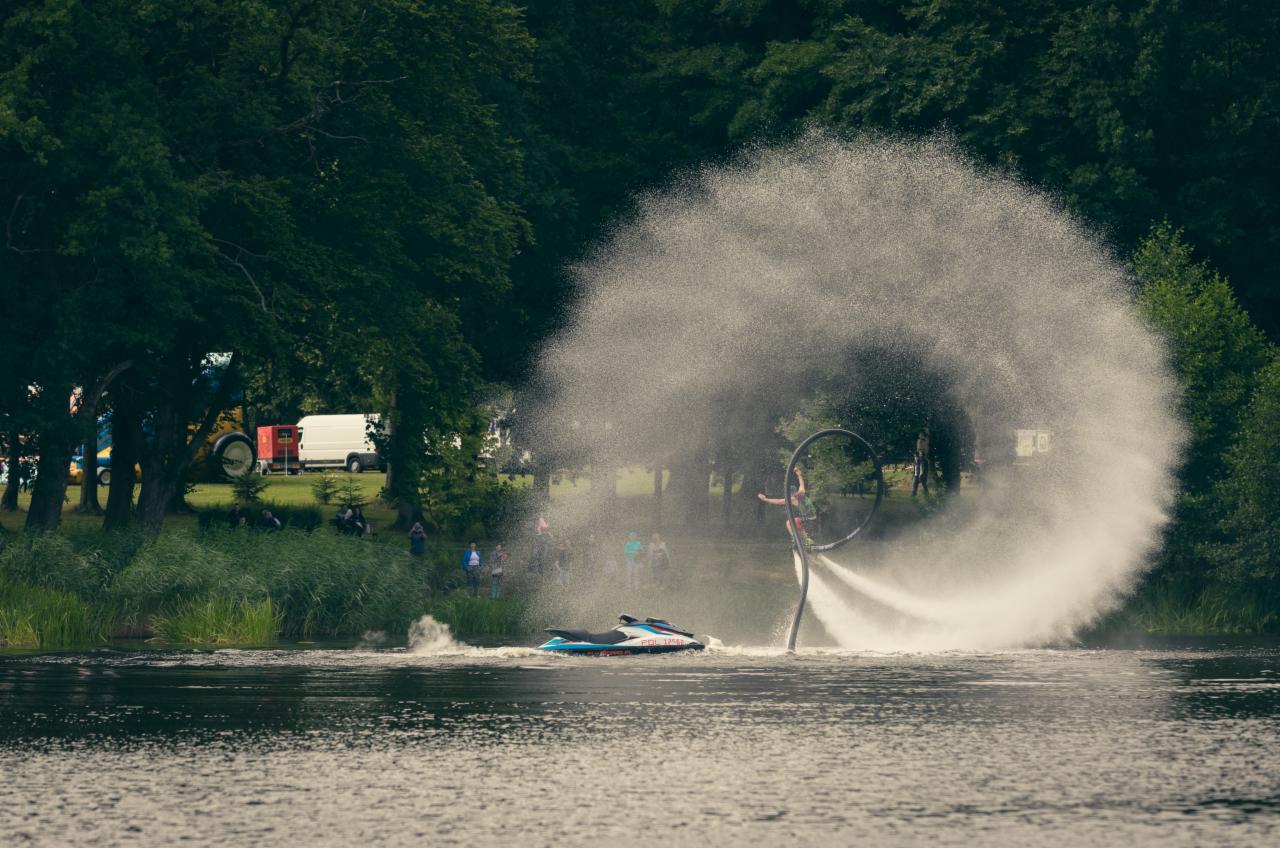 Pokaz Flyboard na jeziorze, strumień wody tworzy efektowny łuk, w tle widoczni widzowie i zieleń drzew.