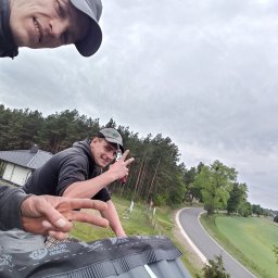 Usługi ogólnobudowlane Arkadiusz Klaman - Dwóch dekarzy na dachu, jeden z logo firmy na bluzie, drugi pokazuje znak zwycięstwa, widok na okolicę z lasem i drogą.