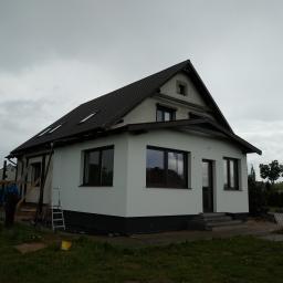 Usługi ogólnobudowlane Arkadiusz Klaman - Dom jednorodzinny z ciemnobrązowym dachem i jasną elewacją, widoczny podczas prac wykończeniowych, z drabina opartą o ścianę i pracownikiem w odzieży roboczej w tle, pochmurne niebo.