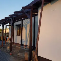 Drewniana konstrukcja zadaszenia tarasu w Bysławiu, widoczna na tle jasnego budynku i kostki brukowej, w trakcie montażu.