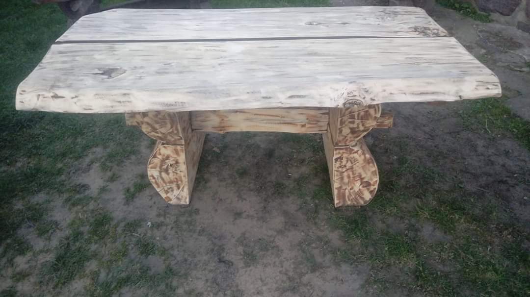 Stół ogrodowy z grubych, bielonych desek i rzeźbionych, bejcowanych nóg, stojący na trawniku.