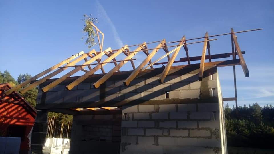 Surowa konstrukcja więźby dachowej z drewnianych belek na murowanym budynku, widoczna zieleń w tle, wczesny etap budowy.