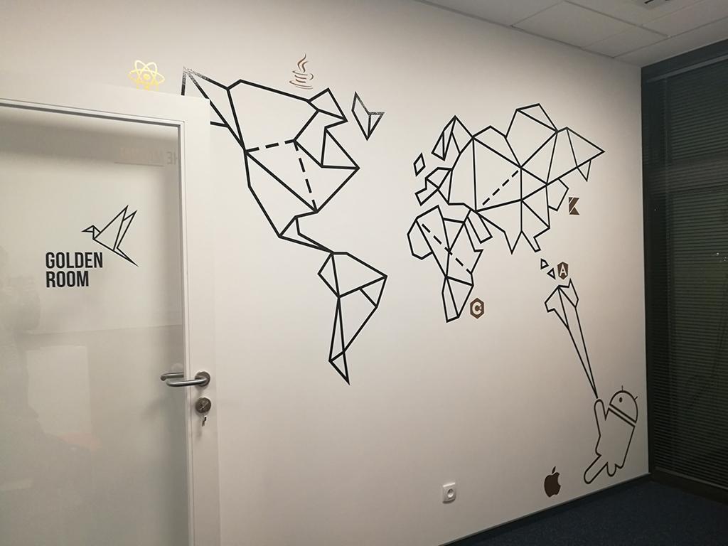 Ściana biura z minimalistyczną, geometryczną mapą świata wykonaną z czarnej taśmy, uzupełnioną ikonami programistycznymi oraz logo Apple i Android, obok drzwi z napisem 'Golden Room' i logo ptaka...