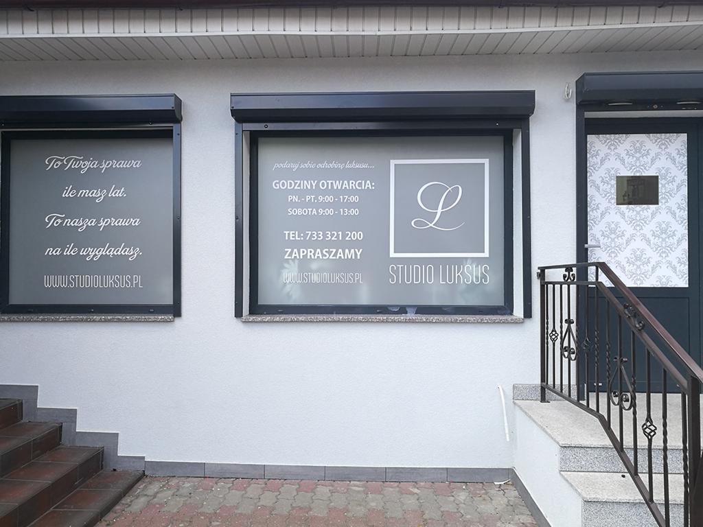 Fasada budynku z dwoma oknami z reklamami i logo Studia Luksus oraz wejściem ze schodami i ozdobnymi wzorami na szklanej części drzwi.