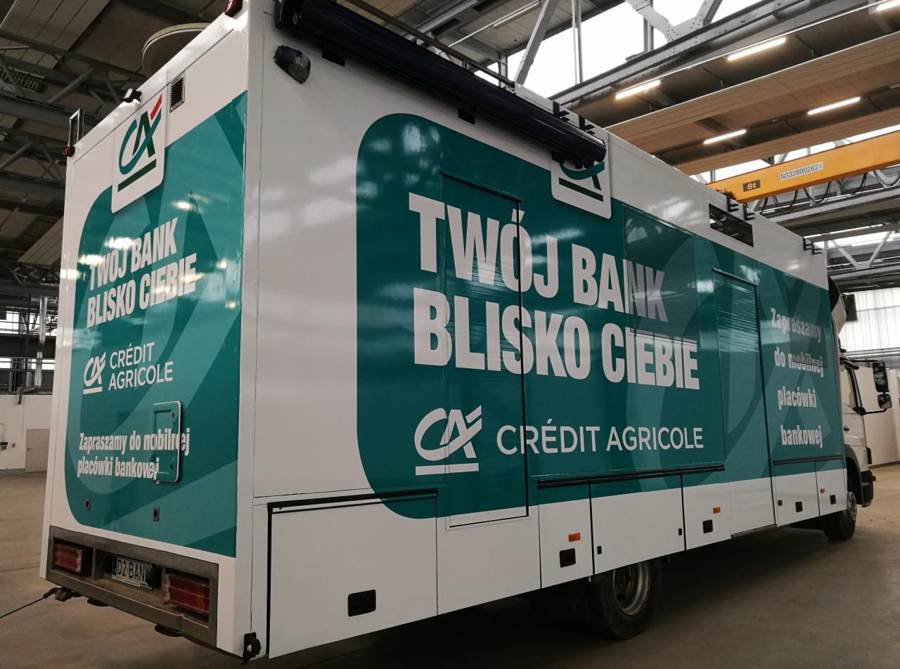 Mobilny oddział banku Crédit Agricole na bazie samochodu ciężarowego z zielono-białą grafiką reklamową, zaparkowany w hali.