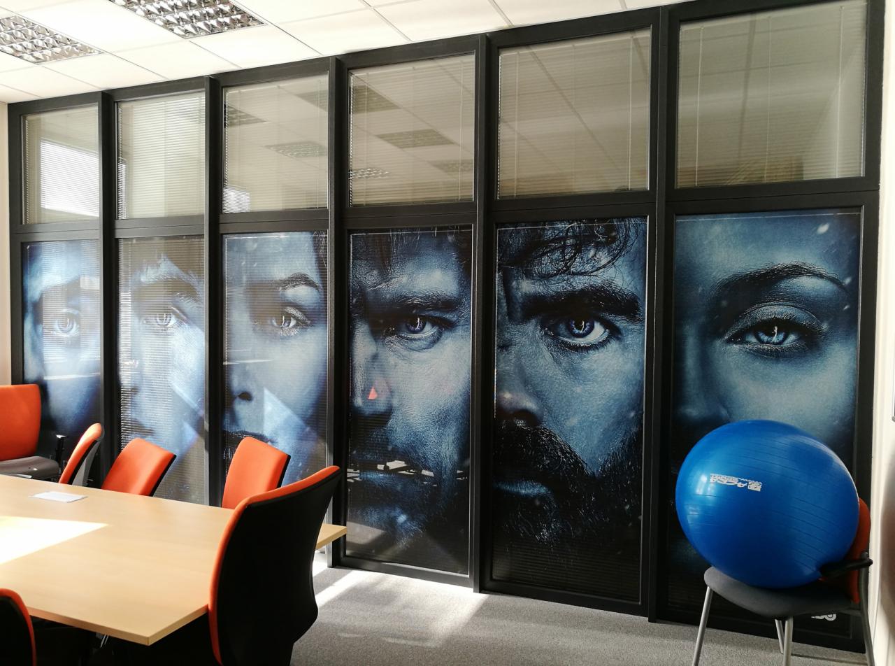 Sala konferencyjna z oklejonymi szybami przedstawiającymi zbliżenia twarzy aktorów z serialu fantasy w odcieniach niebieskiego, pomarańczowe krzesła i niebieska piłka do ćwiczeń.