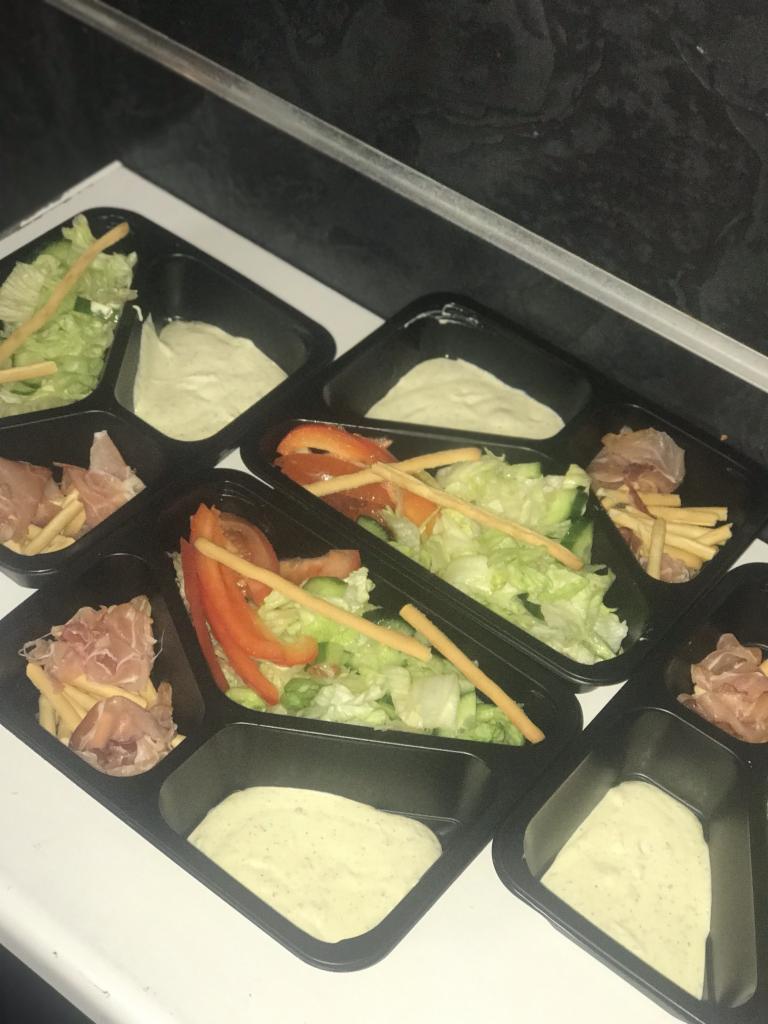 Porcje sałatek z szynką parmeńską, warzywami i sosem w czarnych, plastikowych pojemnikach na białym blacie.
