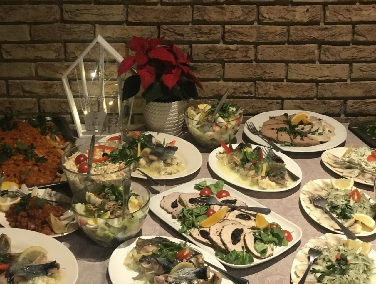 Uroczysty stół z różnorodnymi potrawami: sałatki w szklanych misach, ryba w galarecie, mięsa z dodatkami, dekoracja świąteczna w tle na tle ceglanej ściany.