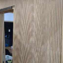 Wykończona ściana z drewnianych paneli pionowych z czarnymi szczelinami, widok na wnętrze przez otwarte drzwi przesuwne.