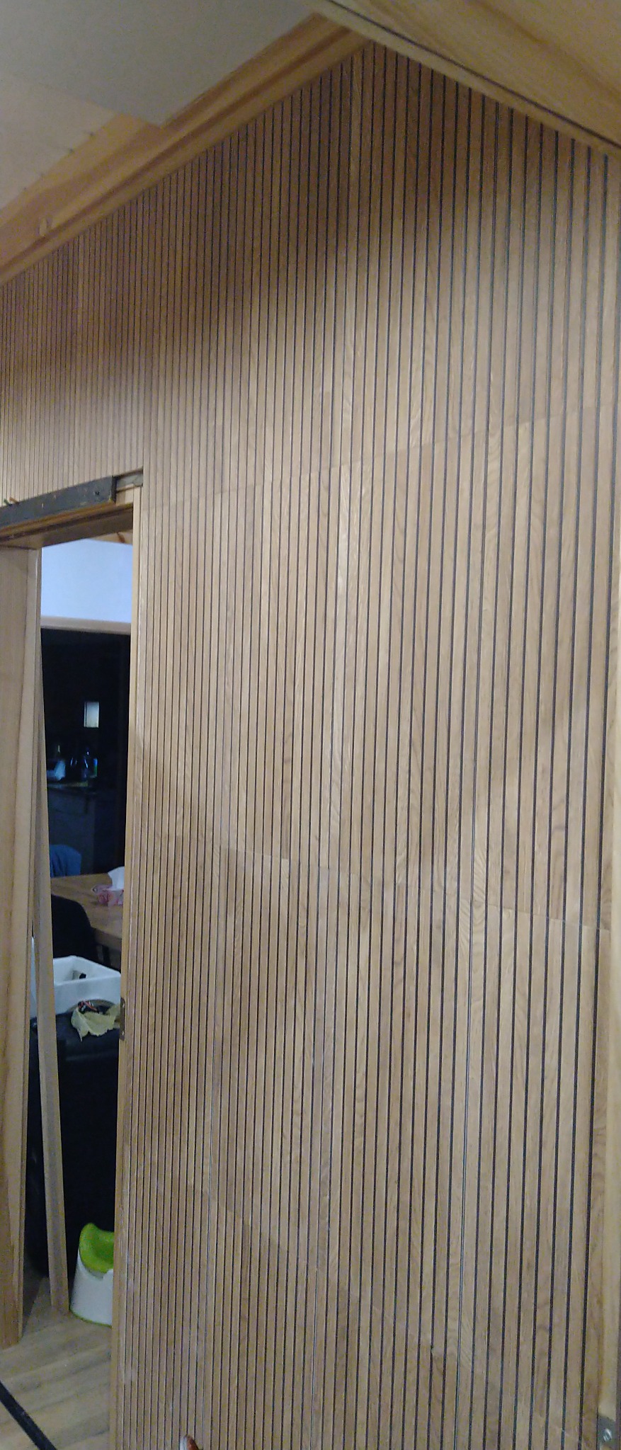 Wykończona ściana z drewnianych paneli pionowych z czarnymi szczelinami, widok na wnętrze przez otwarte drzwi przesuwne.