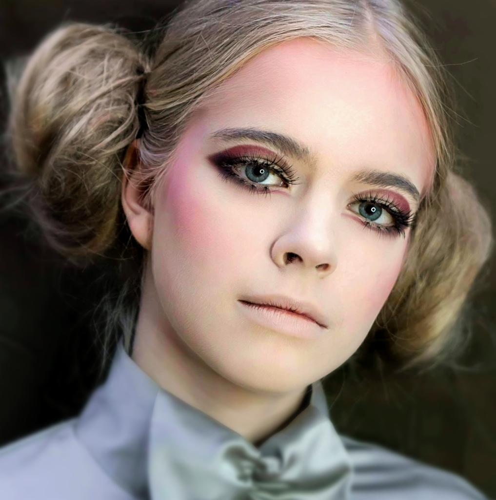 Portret młodej kobiety z intensywnym, burgundowym cieniowaniem powiek, różem na policzkach i włosami upiętymi w dwa koczki, ubrana w satynową bluzkę z kokardą.