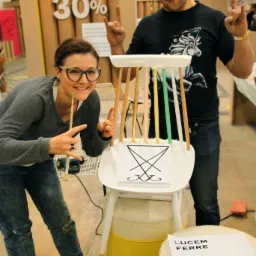 Dwie osoby prezentują pomalowane krzesło z symbolem na oparciu, stojące na żółtej beczce, z podniesionymi rękami w geście rock'n'rolla.