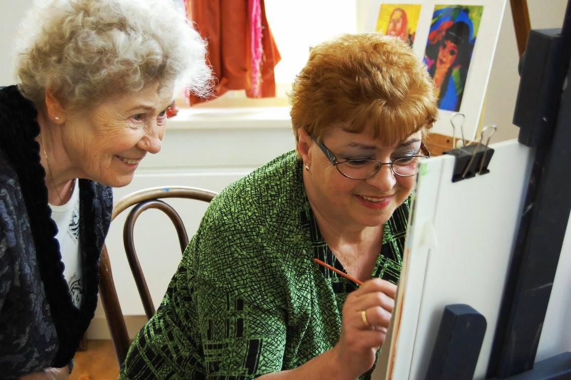 Dwie kobiety, jedna z pędzlem w dłoni, maluje na sztaludze, druga obserwuje z uśmiechem; w tle widoczne obrazy na płótnie.