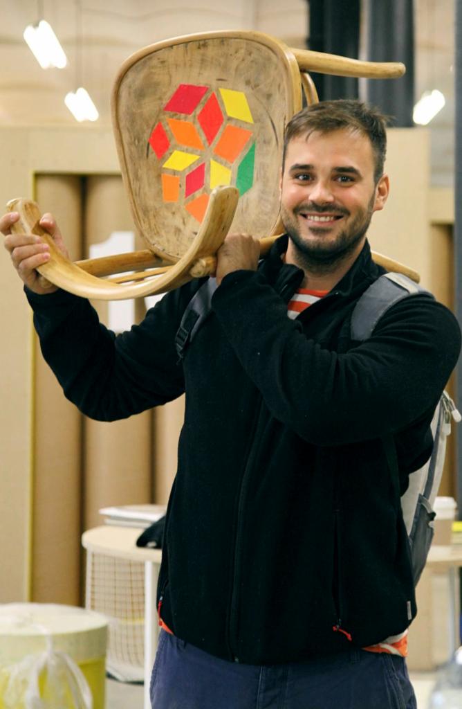 Uśmiechnięty mężczyzna z brodą niesie na ramieniu odnowione krzesło z drewnianym siedziskiem ozdobionym geometrycznym wzorem z kolorowych naklejek w kształcie sześcianów.