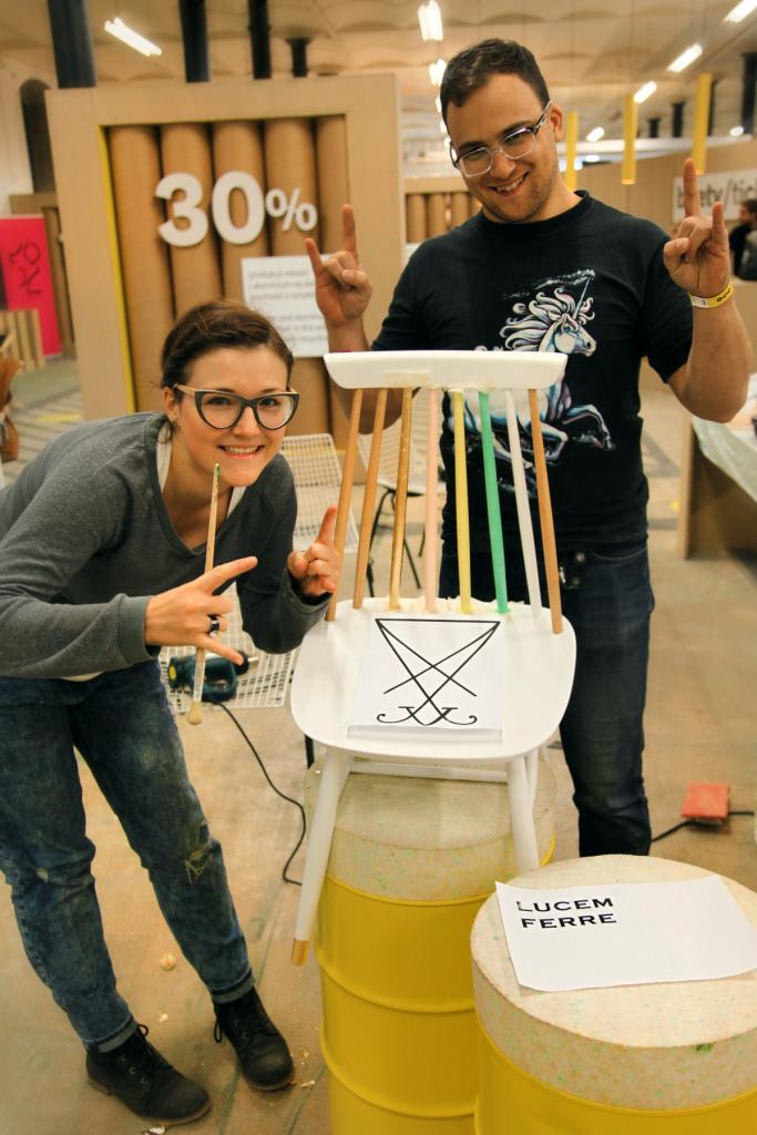 Dwie osoby prezentują pomalowane krzesło z symbolem na oparciu, stojące na żółtej beczce, z podniesionymi rękami w geście rock'n'rolla.