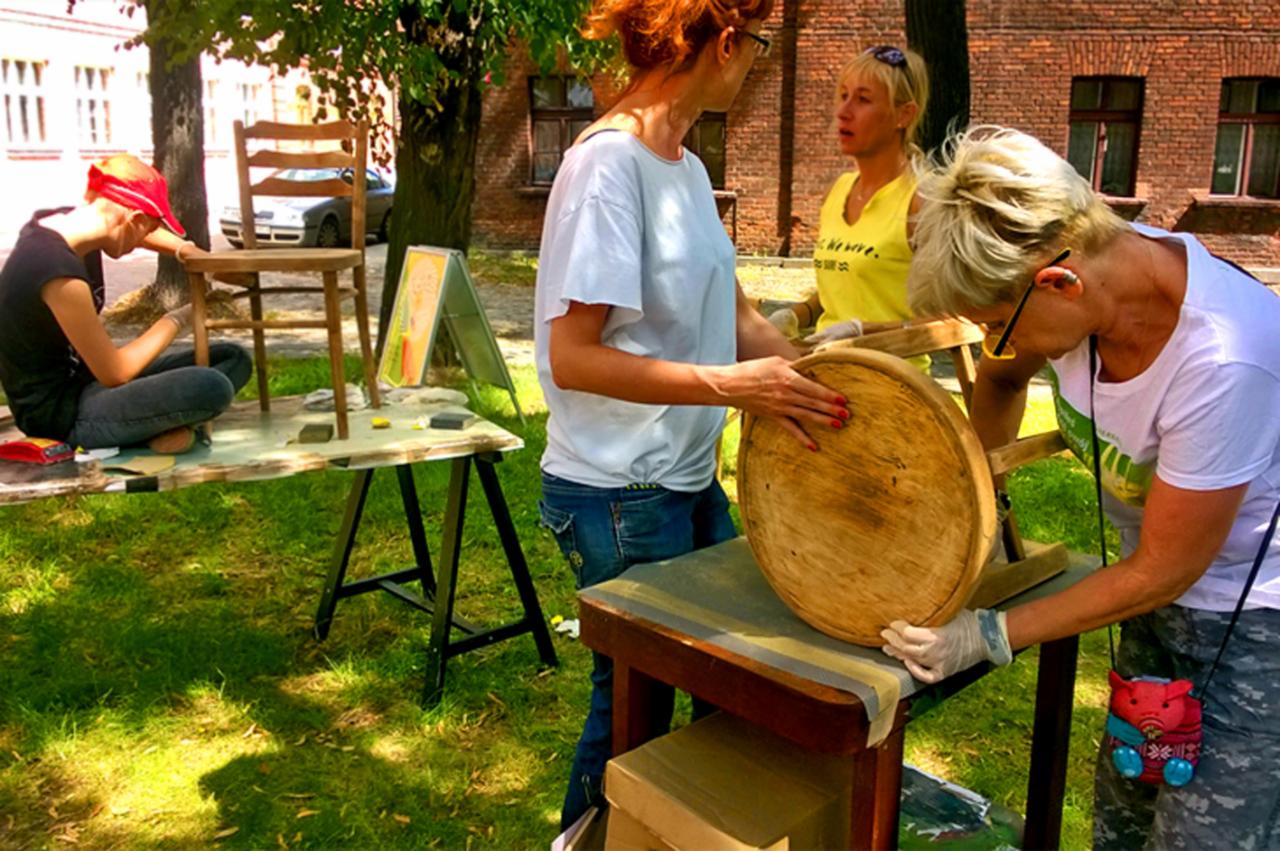 Trzy kobiety pracują na zewnątrz nad renowacją mebli: jedna szlifuje okrągłe siedzisko, druga szlifuje krzesło, trzecia stoi w pobliżu, w tle budynek z czerwonej cegły i drzewa.