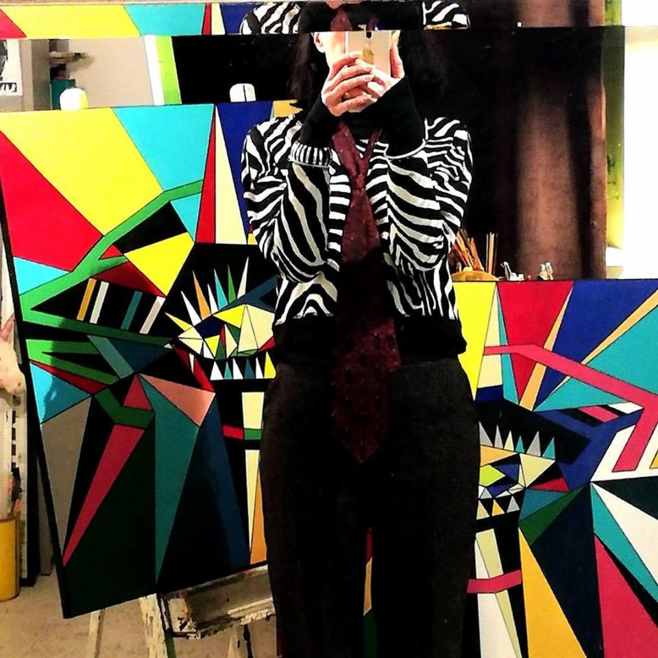 Artystka w bluzie w zebrę robi selfie na tle dwóch geometrycznych obrazów na płótnie w jasnych kolorach, pędzle widoczne w tle.