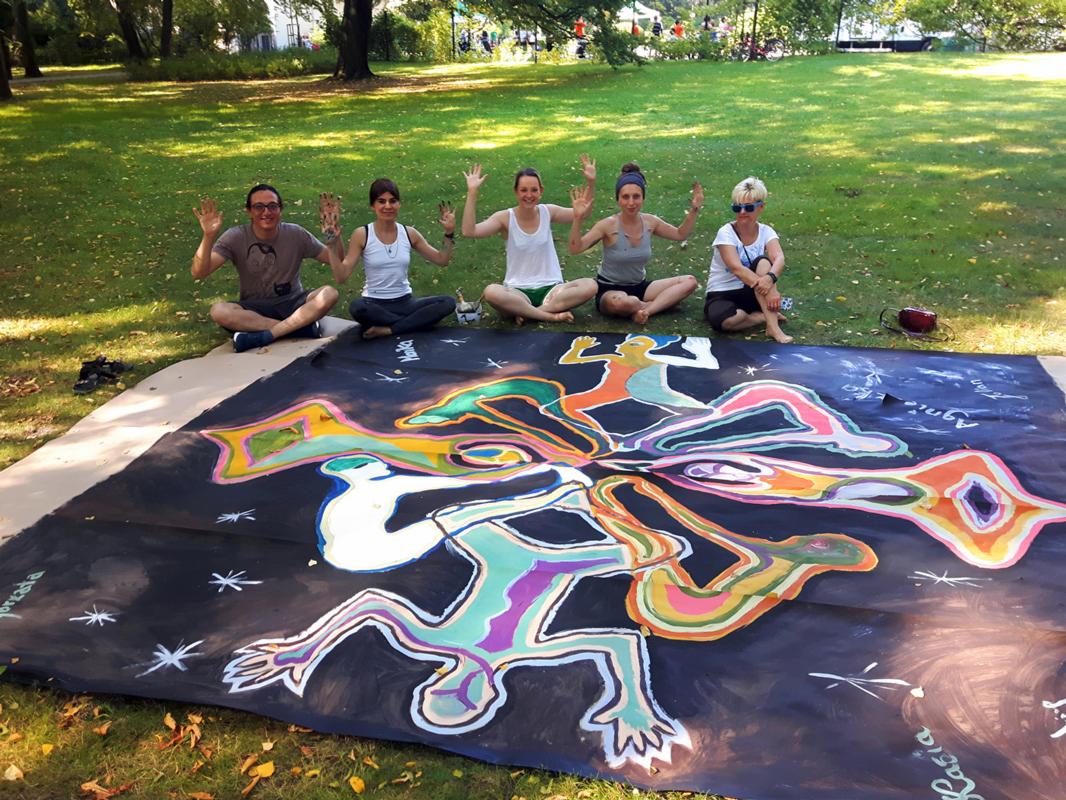 Grupa pięciu osób siedzi na trawie za dużym, czarnym płótnem z abstrakcyjnym, kolorowym malunkiem przedstawiającym figury ludzkie i gwiazdy. Osoby pozują z podniesionymi rękami.