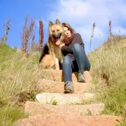 Kobieta siedzi na kamiennych schodach z psem rasy owczarek niemiecki na tle suchej trawy i błękitnego nieba.