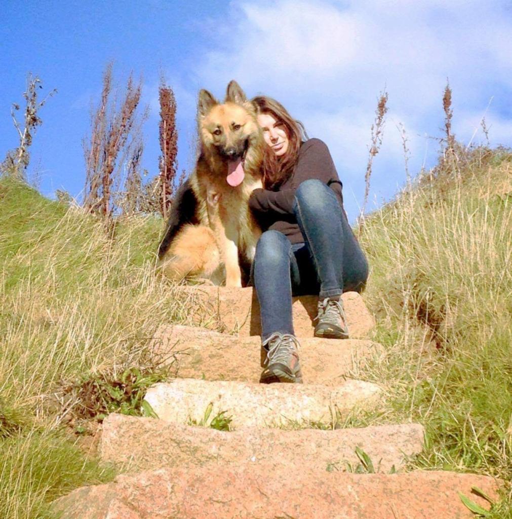 Kobieta siedzi na kamiennych schodach z psem rasy owczarek niemiecki na tle suchej trawy i błękitnego nieba.
