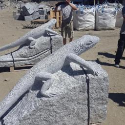 Kopalnia Granitu Górbeh - Dwie rzeźby jaszczurek z szarego granitu na paletach, z widocznymi detalami łusek i pazurów, na tle placu z blokami kamiennymi i pracownikami.