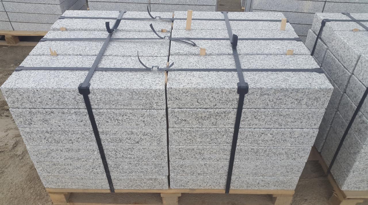 Dwa rzędy szarych, drobnoziarnistych elementów na palecie, spięte czarnymi taśmami, z widocznymi drewnianymi klinami stabilizującymi.