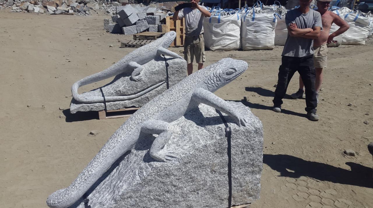 Dwie rzeźby jaszczurek z szarego granitu na paletach, z widocznymi detalami łusek i pazurów, na tle placu z blokami kamiennymi i pracownikami.