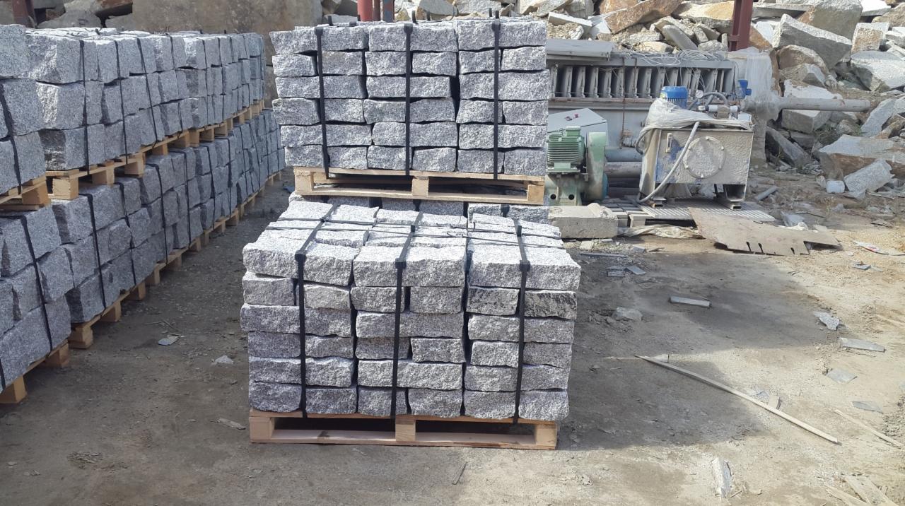 Szare, granitowe kostki ułożone w rzędach na drewnianych paletach, przygotowane do transportu, z widocznymi elementami maszyn do obróbki kamienia w tle.