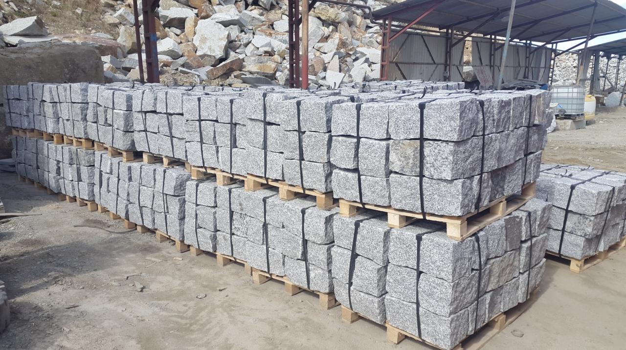 Ułożone palety z szarą kostką, zabezpieczone czarną taśmą, na tle placu z materiałami budowlanymi i konstrukcją zadaszenia.