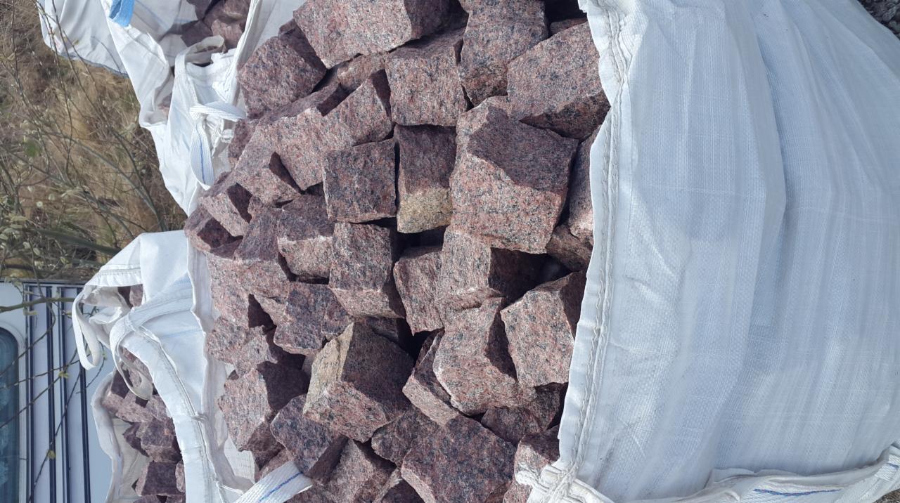 Czerwono-szare, surowe, granitowe sześciany w worku typu big-bag, widoczne tło z zaroślami.