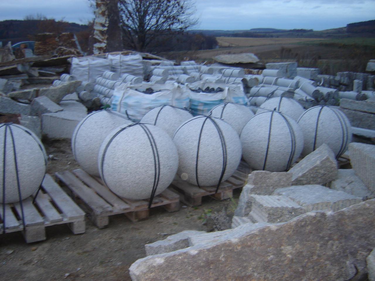 Magazyn kostki granitowej: Granitowe kule na paletach, kostka w workach i bloczki w tle, pochmurne niebo.