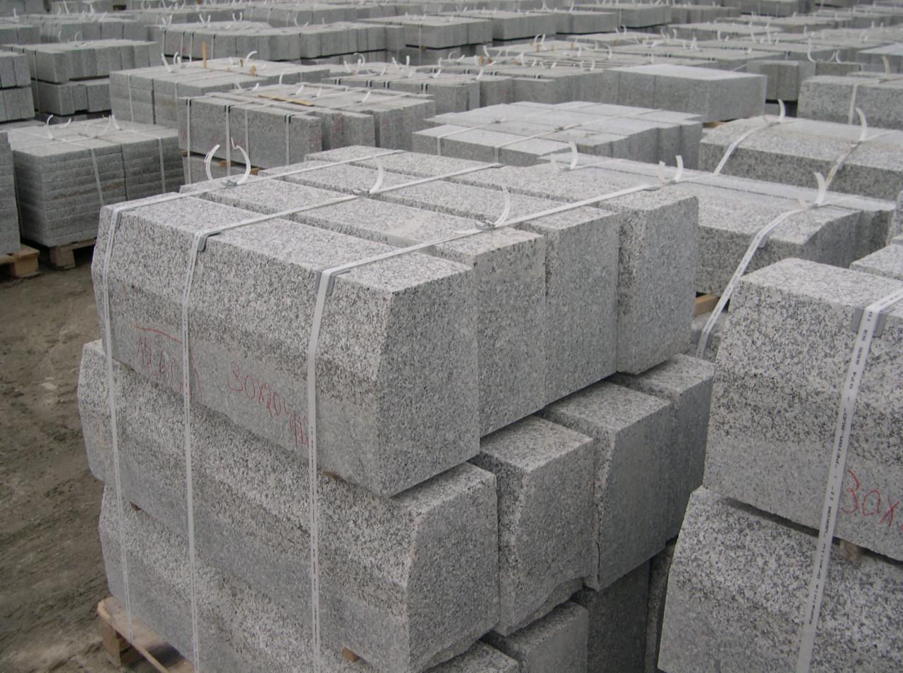 Sztaple szarej kostki granitowej, spięte białymi taśmami na paletach, z widocznymi czerwonymi napisami rozmiarowymi na niektórych kostkach, ułożone w magazynie zewnętrznym.