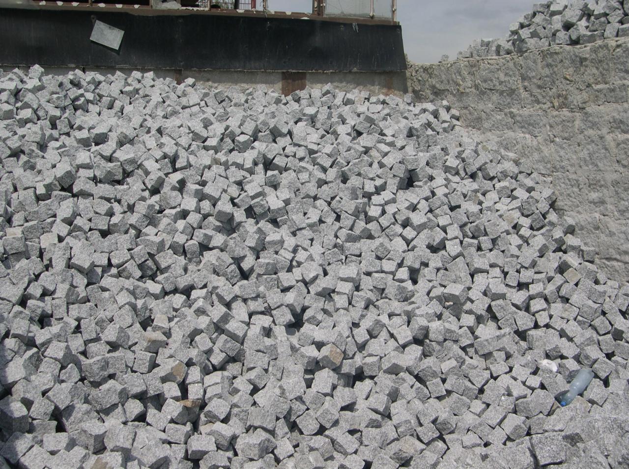 Stos szarej, surowej kostki granitowej o nieregularnych kształtach, na tle kamieniołomu i częściowo widocznego rusztowania.