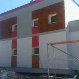 Nowoczesny budynek z częściowo wykończoną elewacją: szare ściany z drewnopodobnymi panelami i czerwonymi akcentami, widoczne rusztowanie i gruz.