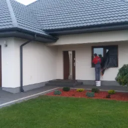 Pracownik w czerwonej bluzie maluje elewację domu jednorodzinnego na biało, stojąc na plastikowym wiadrze, okno zabezpieczone folią, widoczna antena satelitarna i zadbany ogród.