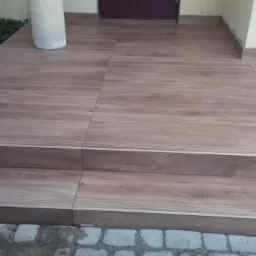 Wyłożone płytkami imitującymi drewno schody wejściowe do domu, z widoczną kolumną i fragmentem elewacji.