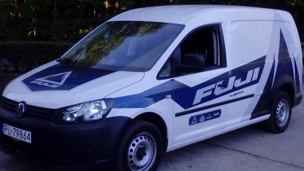 Biały samochód dostawczy z niebieskimi elementami graficznymi i logotypem Fuji Bikes, zaparkowany na zewnątrz w pochmurny dzień.