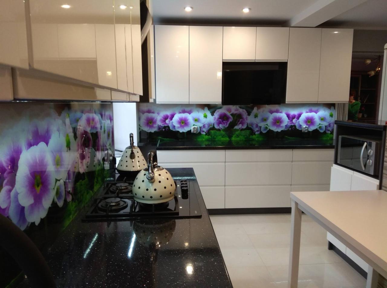 Nowoczesna kuchnia z białymi szafkami i czarnym blatem, na którym stoją dwa czajniki w kropki. Ściana nad blatem ozdobiona szkłem z motywem kwiatowym w odcieniach fioletu i zieleni.