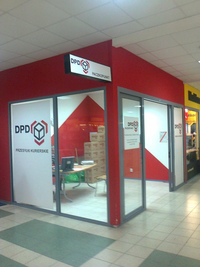 Oklejona witryna punktu kurierskiego DPD z widocznym logo i napisem 'Przesyłki Kurierskie', wnętrze z biurkiem i stosem paczek, czerwono-biała kolorystyka.