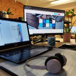 Circinus Training Center Adam Pluciński - Biurko z dwoma monitorami, laptopem z otwartym portfolio firmy szkoleniowej Circinus, słuchawkami i kubkiem. Widoczny fragment ceglanej ściany i rośliny doniczkowe.